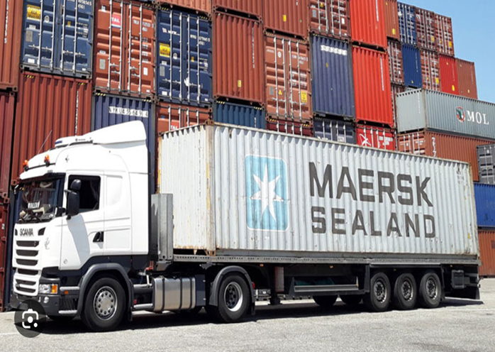 a truck with a container in front of a stack of shipping containers